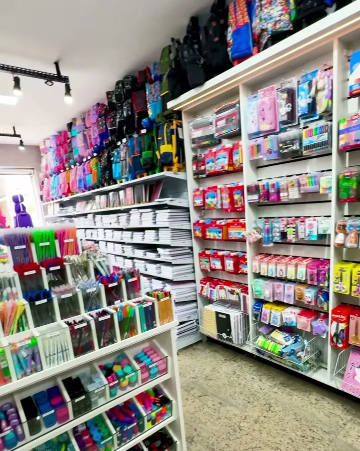 Corredor da loja com prateleiras de cadernos, canetas e estojos até o teto